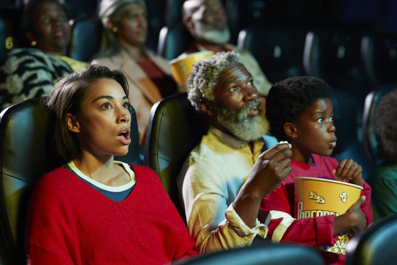 A parent, grandparent, and child holding a bucket of popcorn react with surprise while watching a movie.