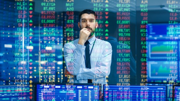 A man in a shirt and tie watches stock tickers and various screens. 