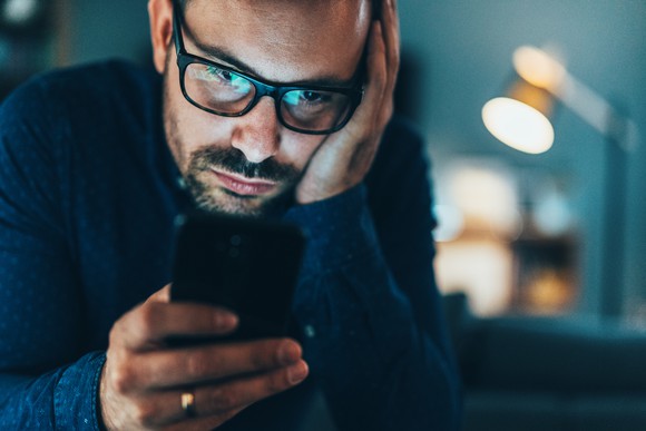 A serious young man looking at his phone with his other hand on the side of his face.