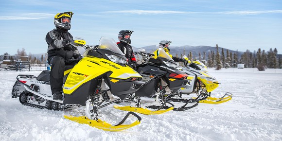 Three riders on Ski-Doo snowmobiles in a snowy landscape, with pine forest, hills, and a blue sky in the background.