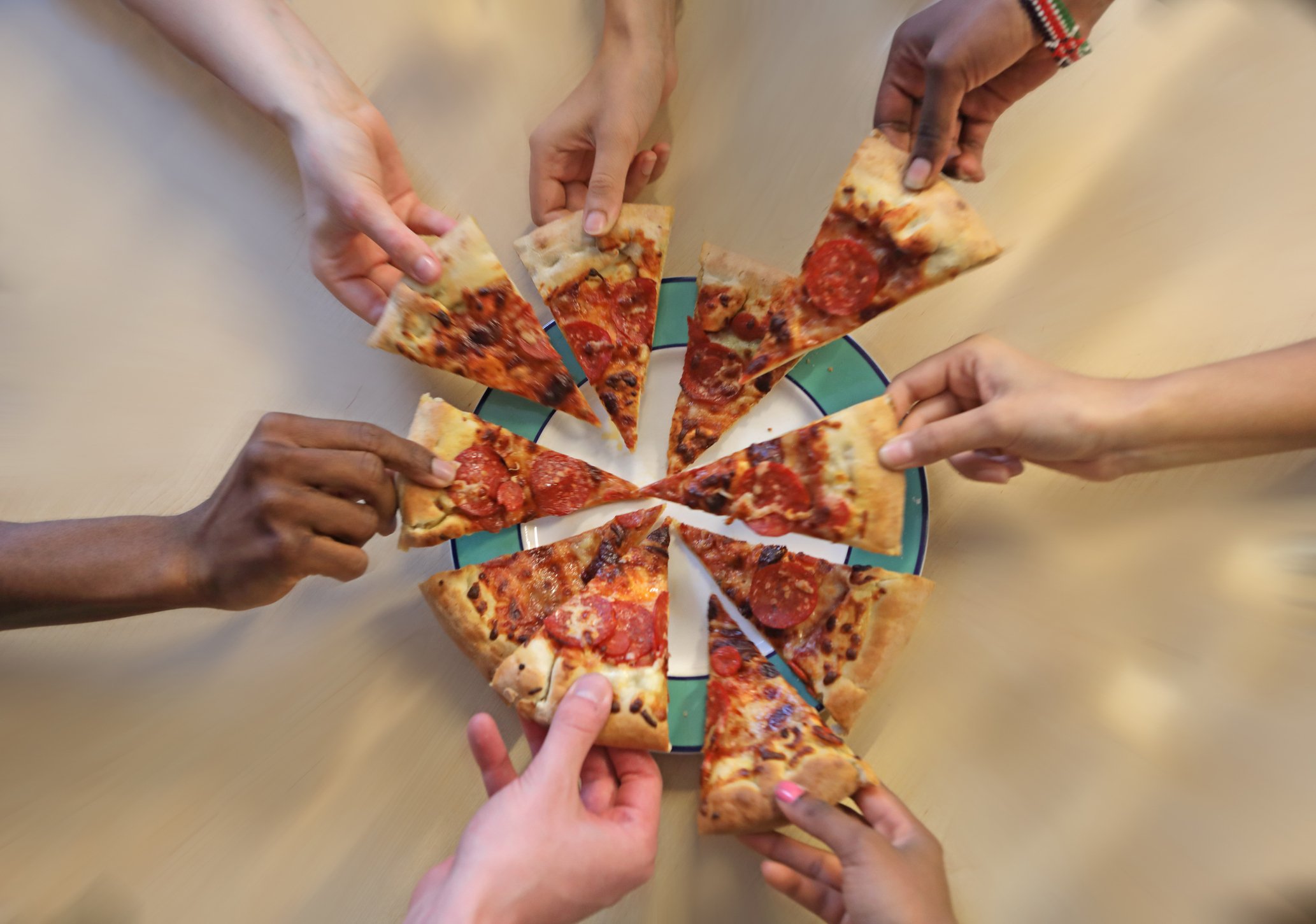 Seven hands taking slices of pizza.