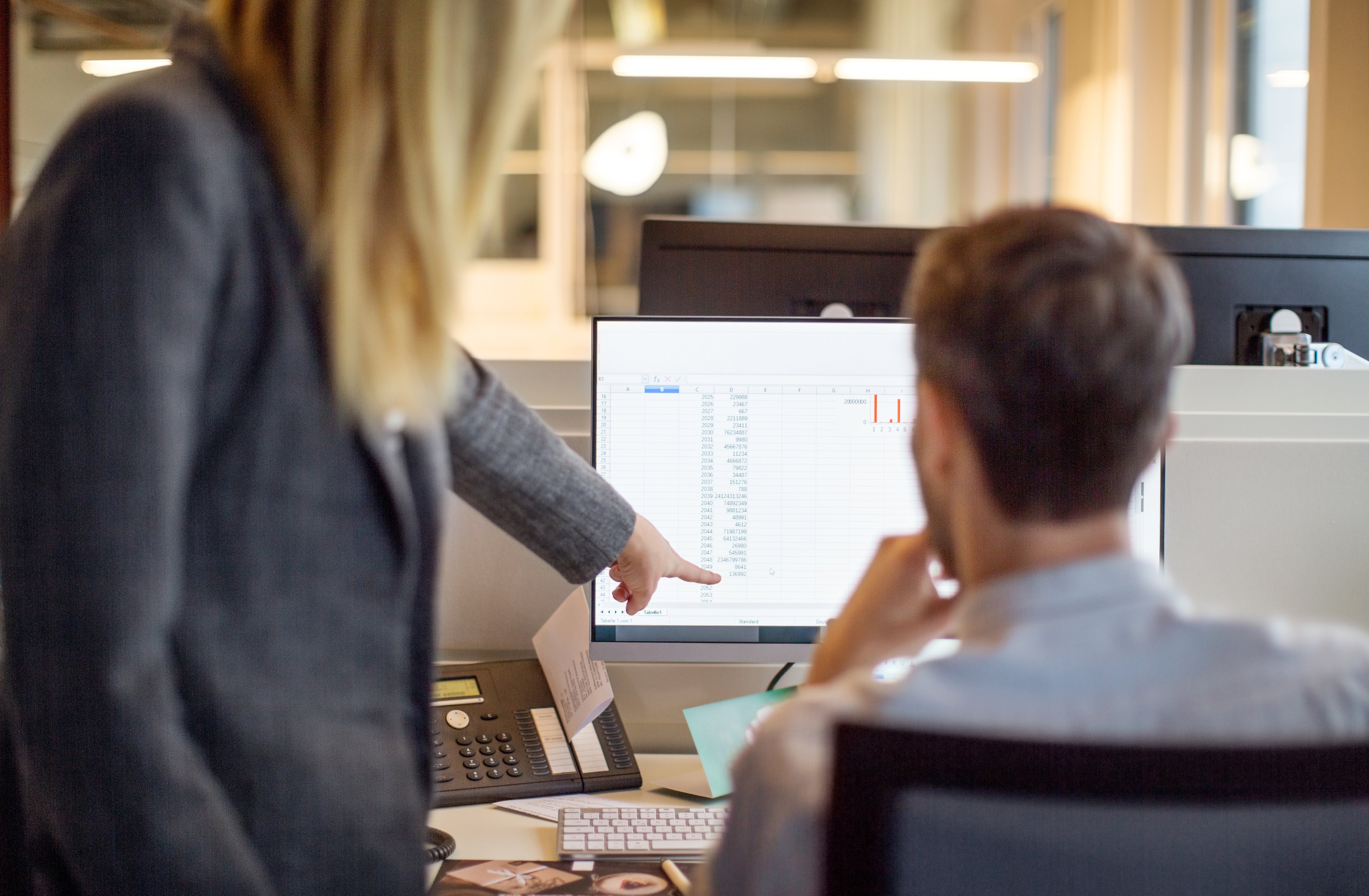 A woman points to a line on a spreadsheet while looking at a computer monitor with a man.