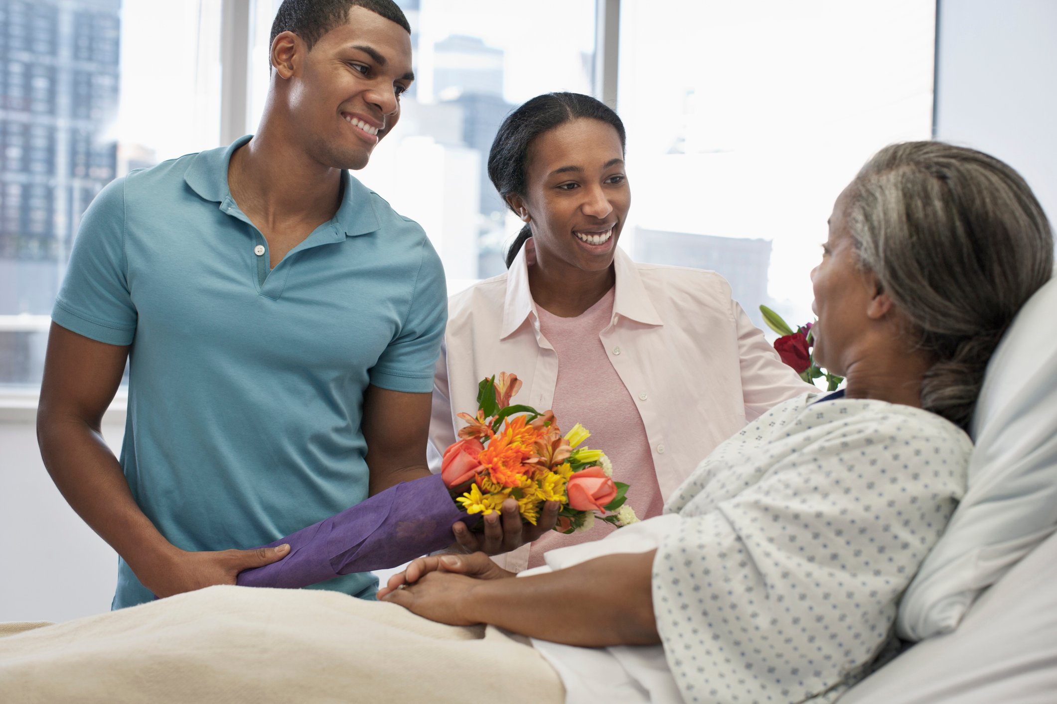 Two young adults bringing flowers to an older person lying in a hospital bed.