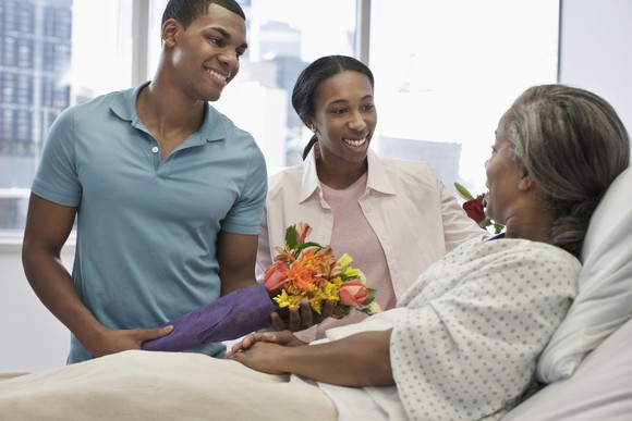 Two young adults bringing flowers to an older person lying in a hospital bed.
