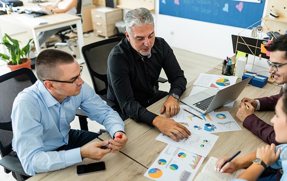 Four coworkers sit around a table, considering several printouts of sales data.