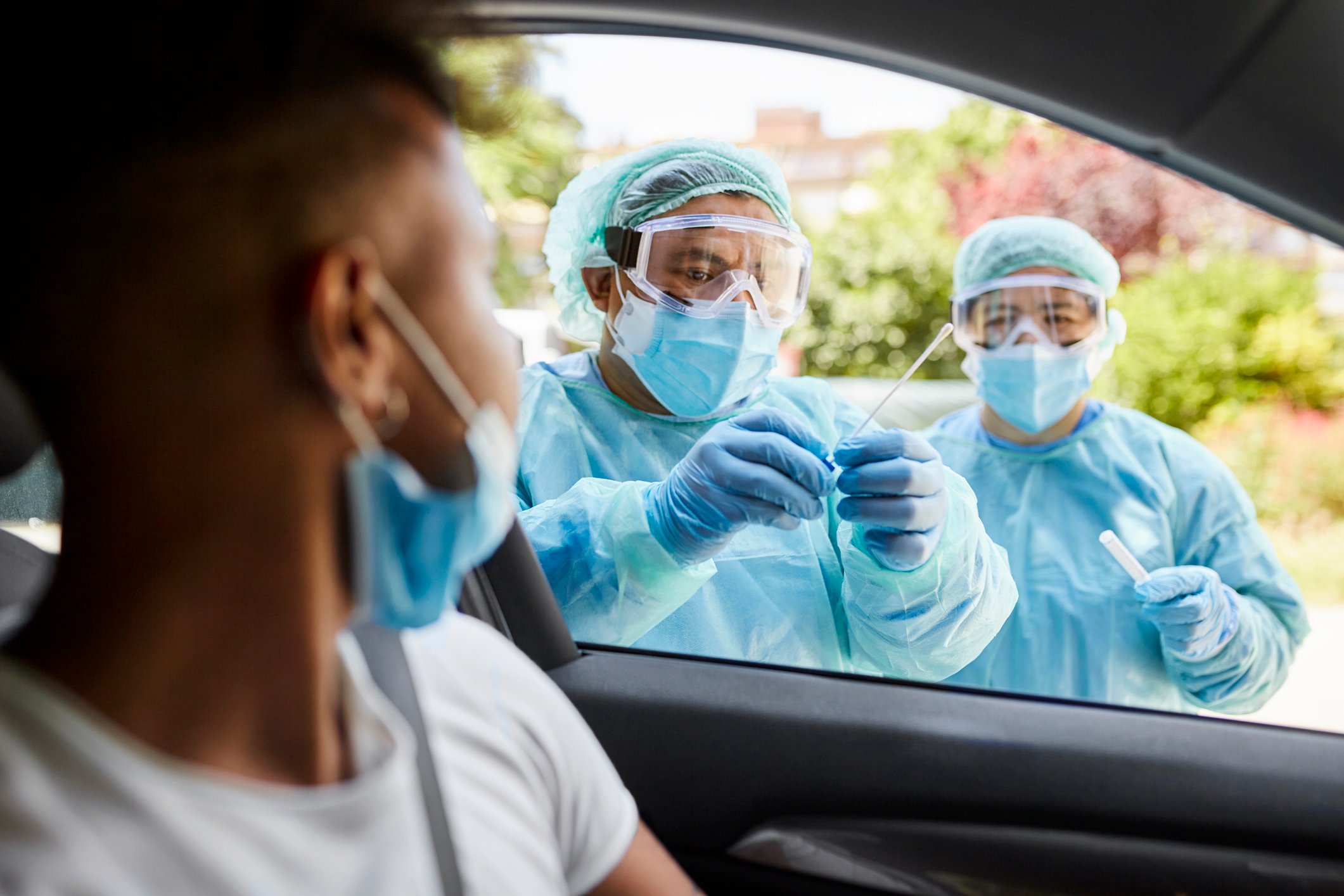 Two healthcare workers in protective gear get a sample from someone at a drive-thru testing site.