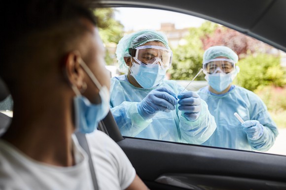 Two healthcare workers in protective gear get a sample from someone at a drive-thru testing site.
