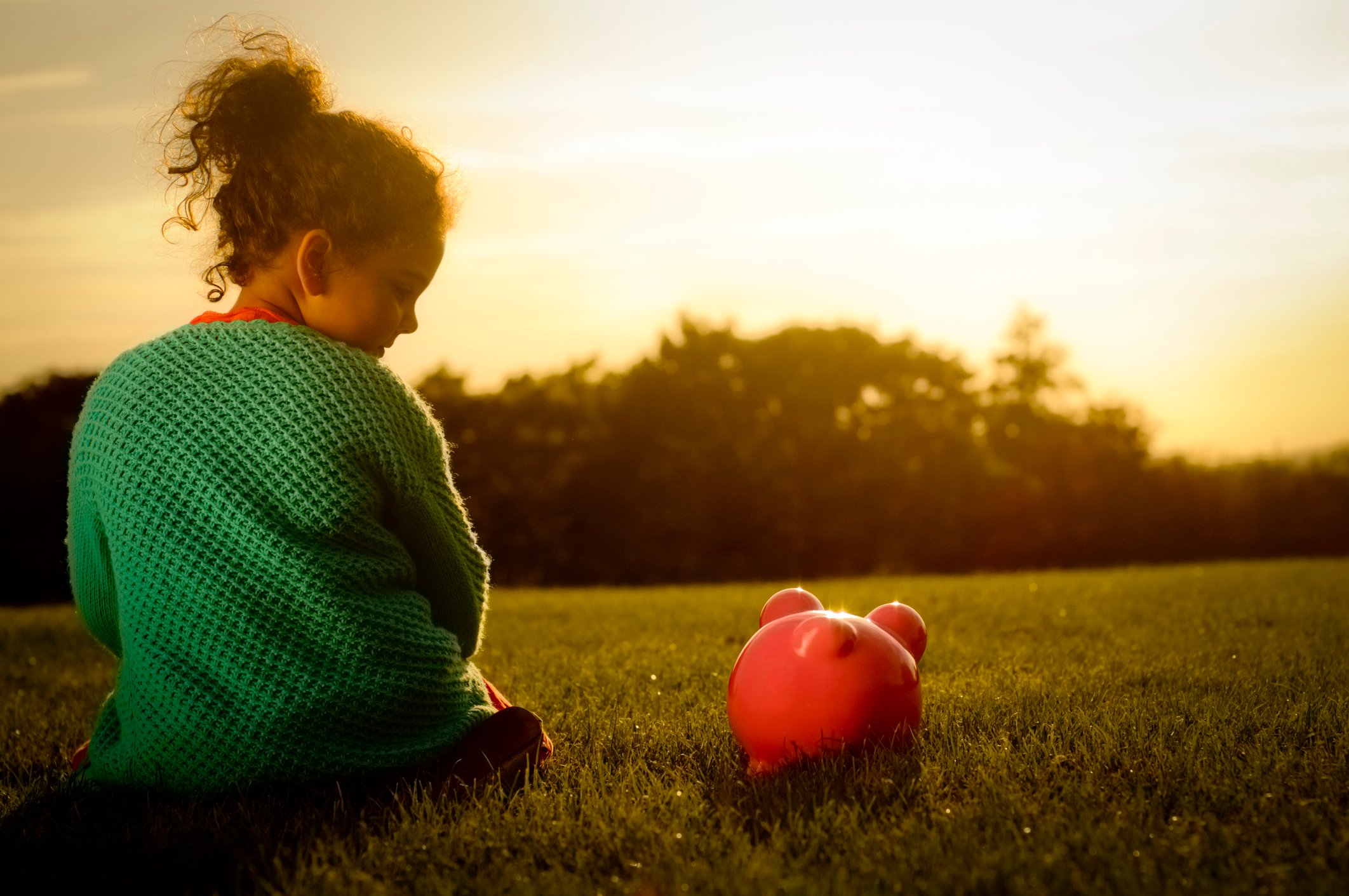 A girl looks down at her piggy bank while the sun sets in the background. 