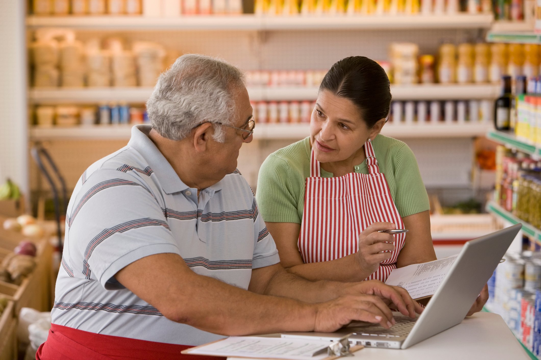 Two business owners discuss information they're reviewing on a laptop and a clipboard.