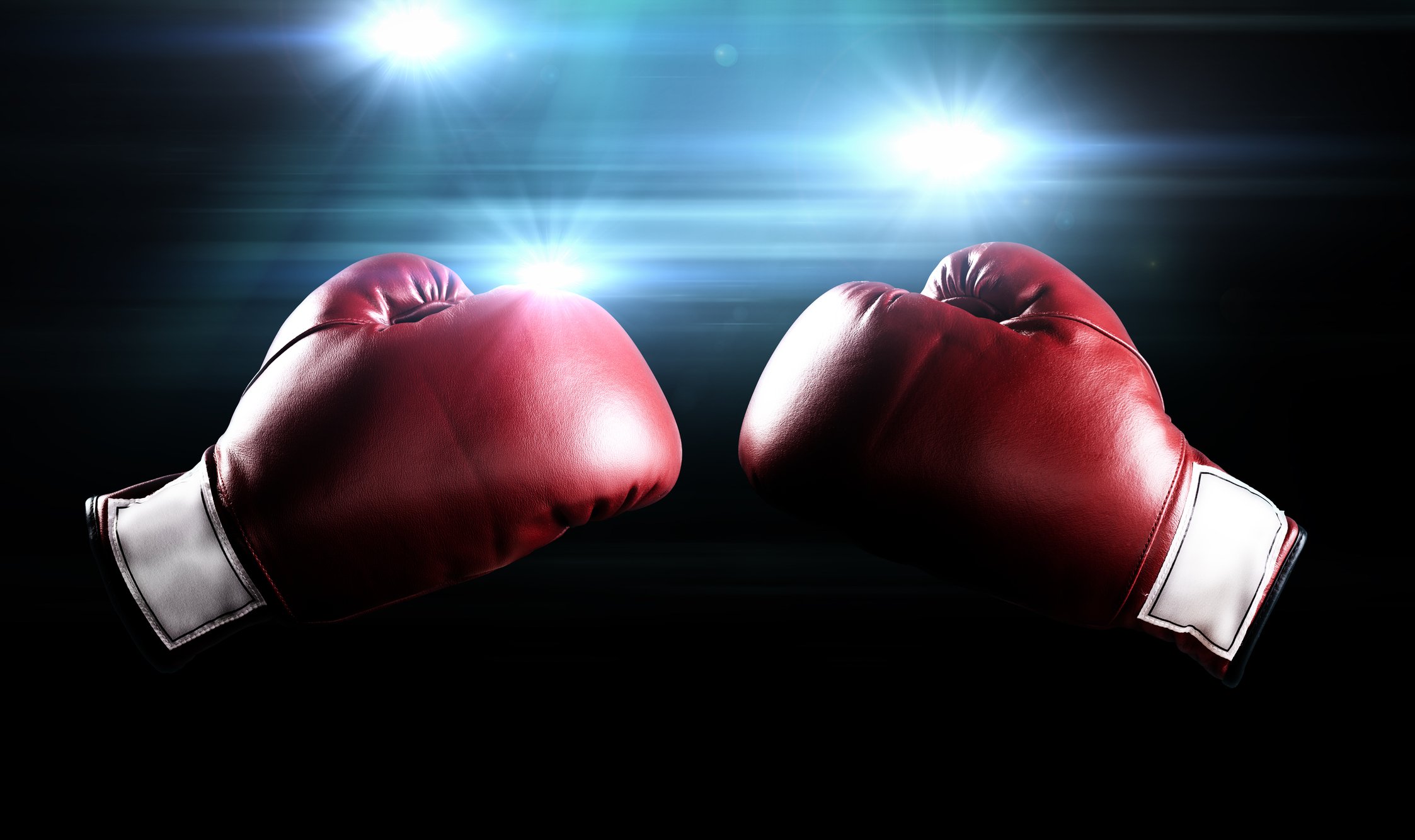 Two red boxing gloves facing each other with lights shining in the background.