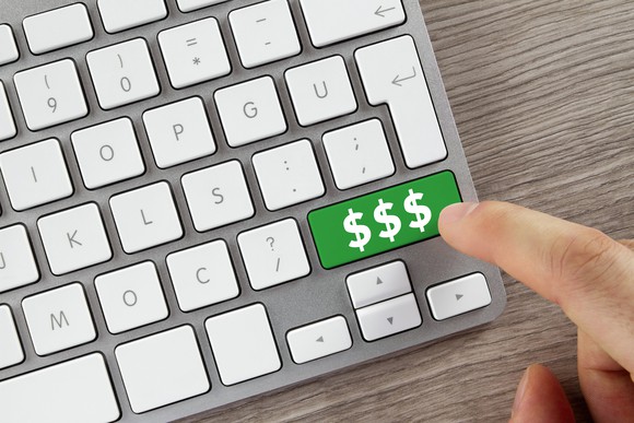 An index finger on top of a green button on a computer keyboard with three dollar signs on the button.