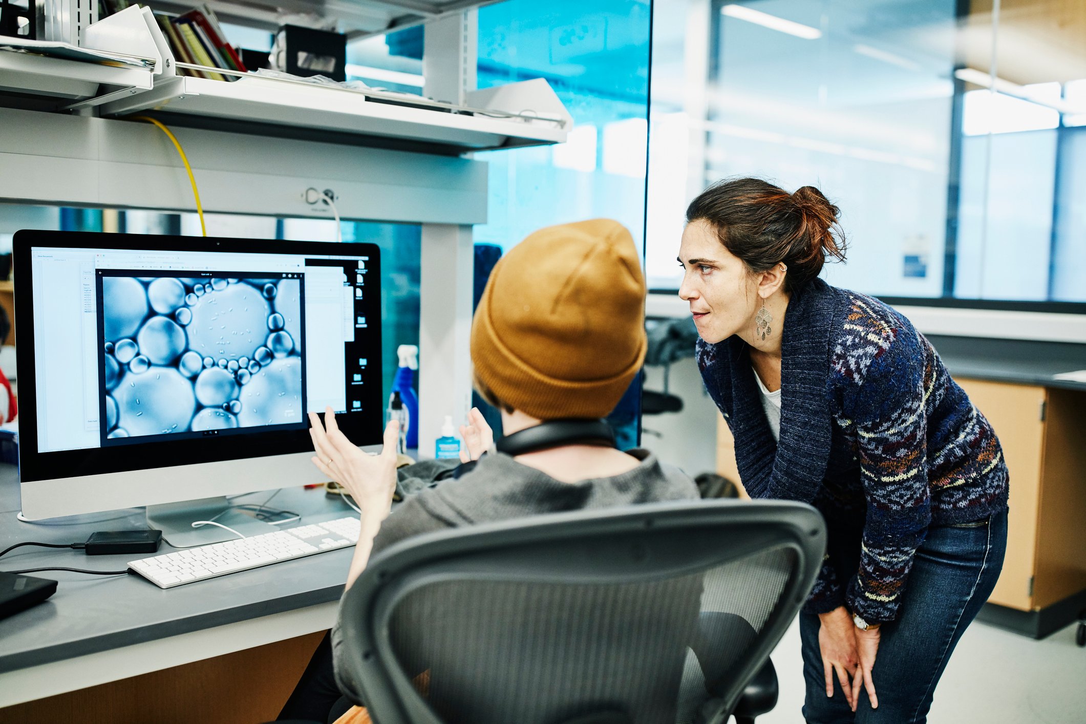 A woman looks with another person at a computer monitor in a lab setting.