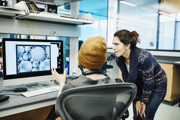 A woman looks with another person at a computer monitor in a lab setting.