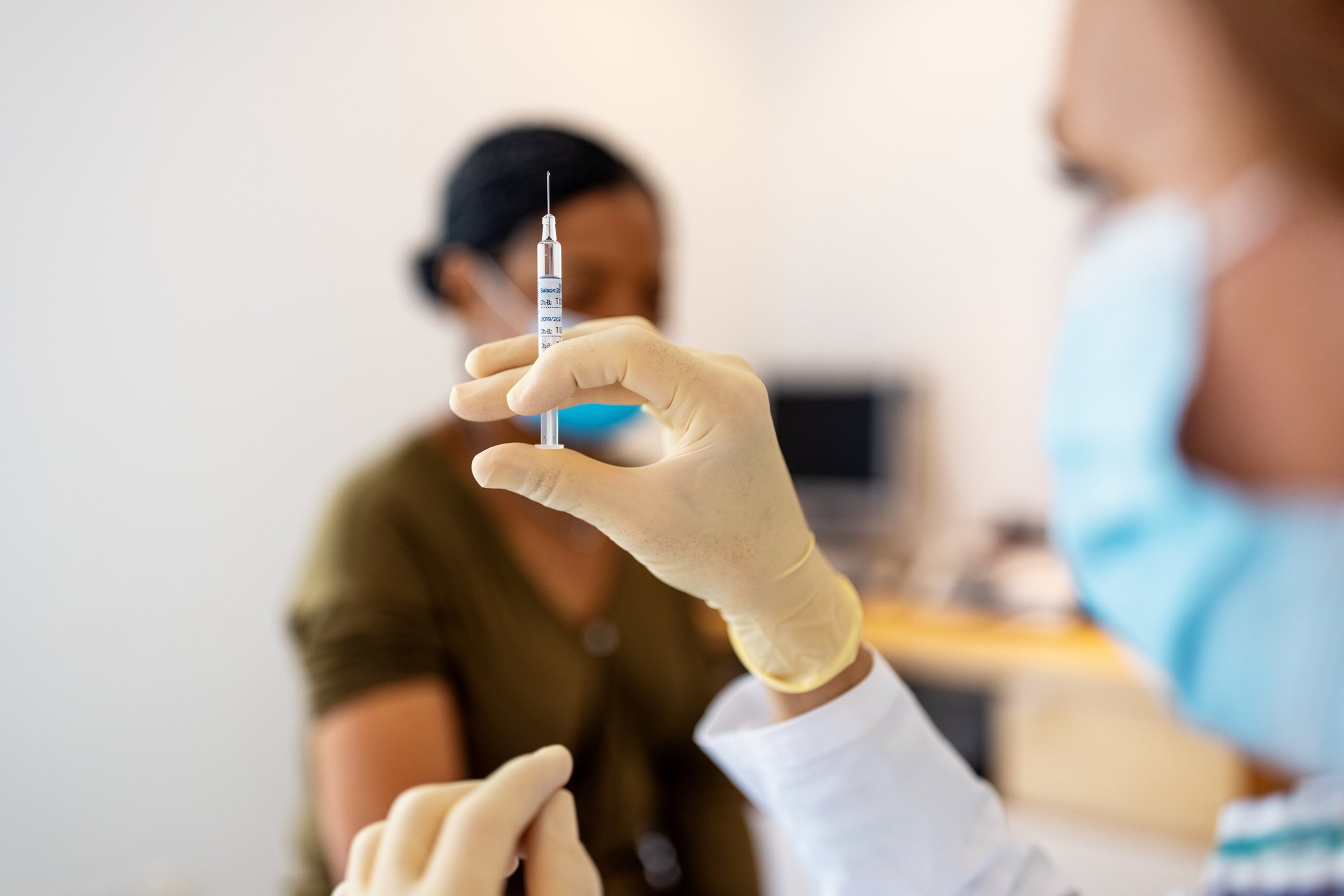 A nurse holds a hypodermic needle as a patient waits in the background. 