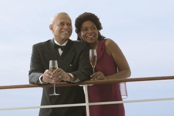 A senior couple in formal wear holding champagne glasses while aboard a cruise ship. 