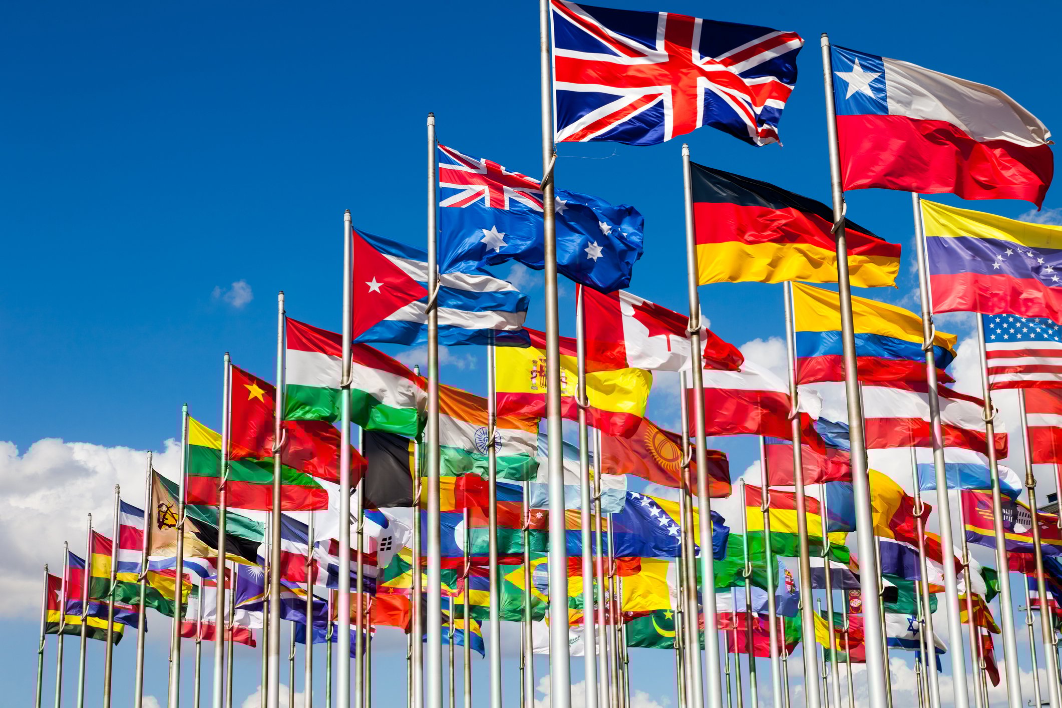 Flags of many nations flying on flagpoles against a backdrop of blue sky.