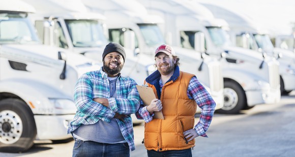 Two smiling people with five semi-trailer trucks in the background.