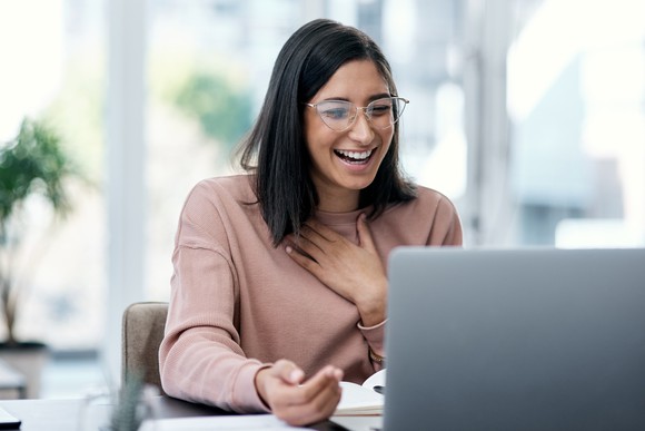 A woman smiling, hand on her chest, pleased at what she is reading on a laptop. 