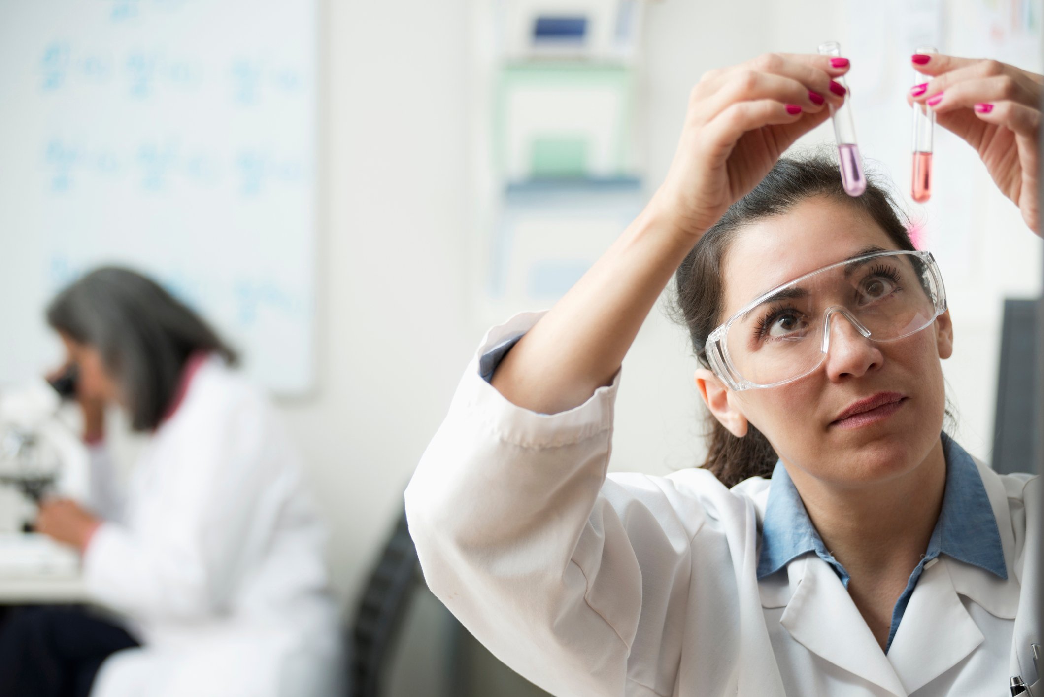 A scientist examines two test tubes as another scientist in the background looks into a microscope.