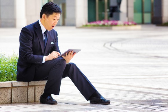A man in a suit recoils at his tablet sitting outside. 