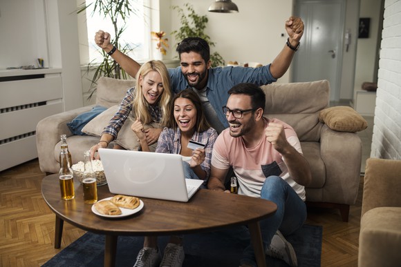 A group of young people gathered around a living room table and reacting triumphantly to something on a laptop PC.