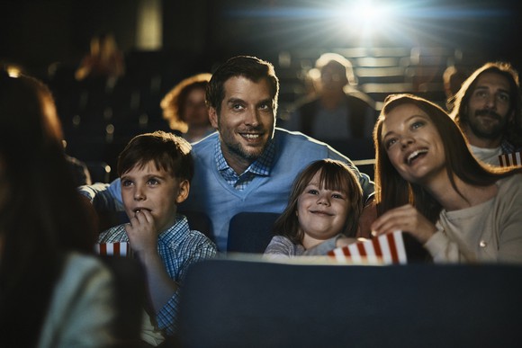 Two adults and two children eat popcorn and watch a movie in a theater.