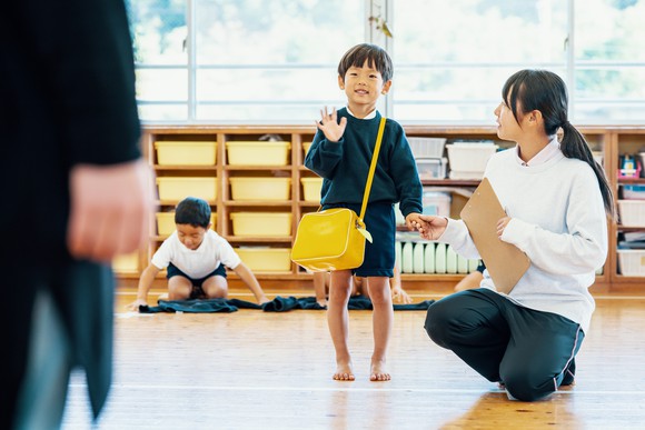 A young boy in a classroom, holding hands with an adult and waving.