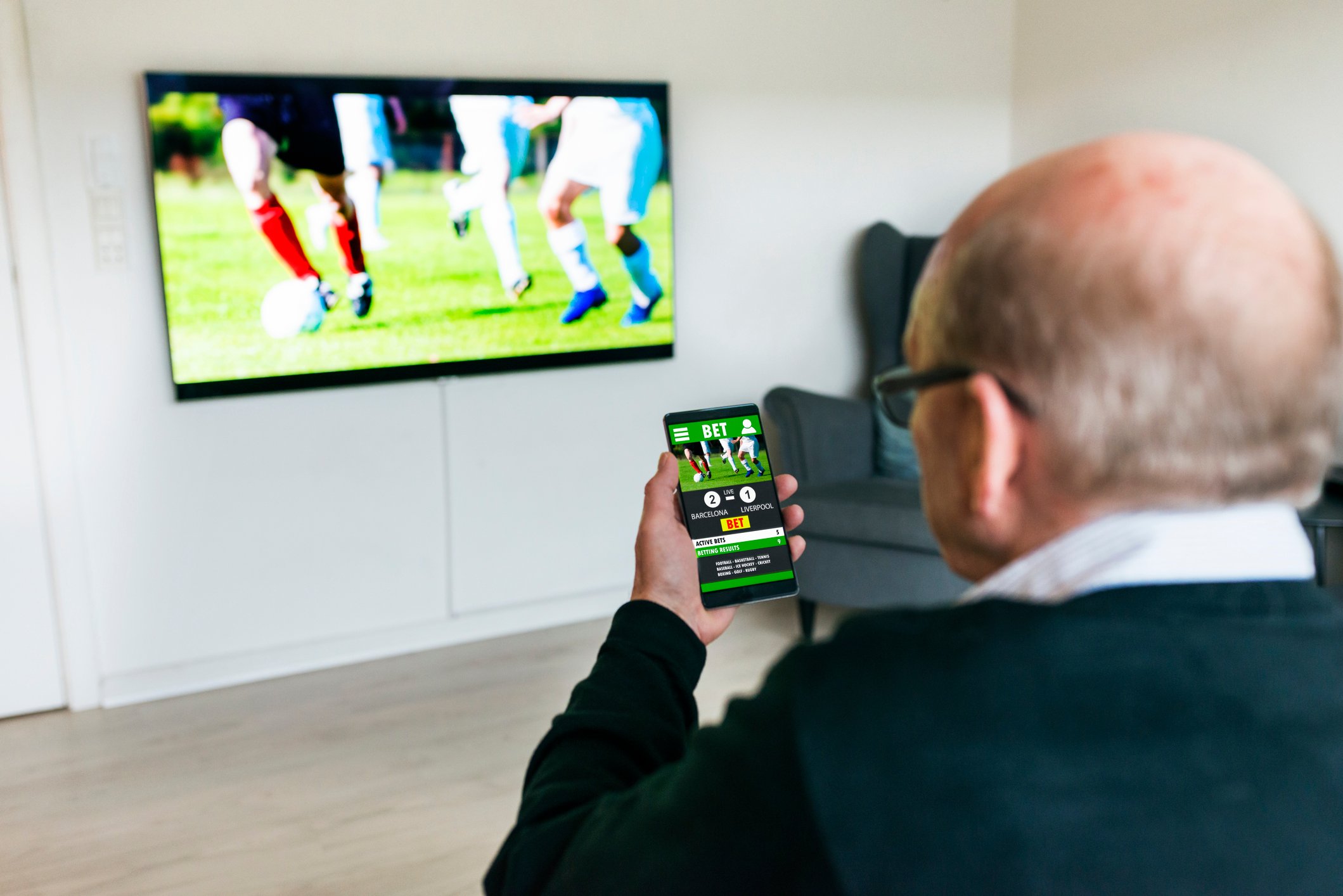 A person watching a soccer match on a large TV while making a bet on a mobile device.