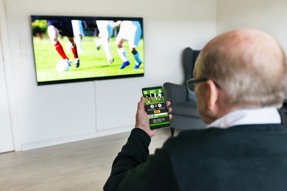 A person watching a soccer match on a large TV while making a bet on a mobile device.