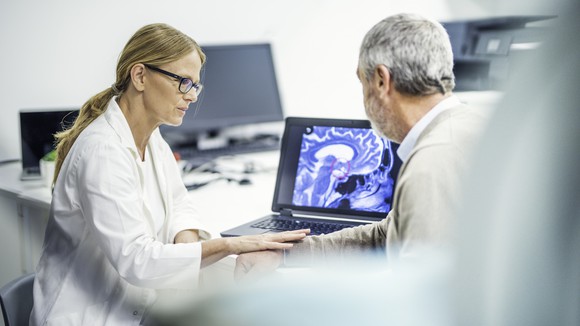 A medical professional shows an image of a brain scan on a laptop to another person while touching their arm.