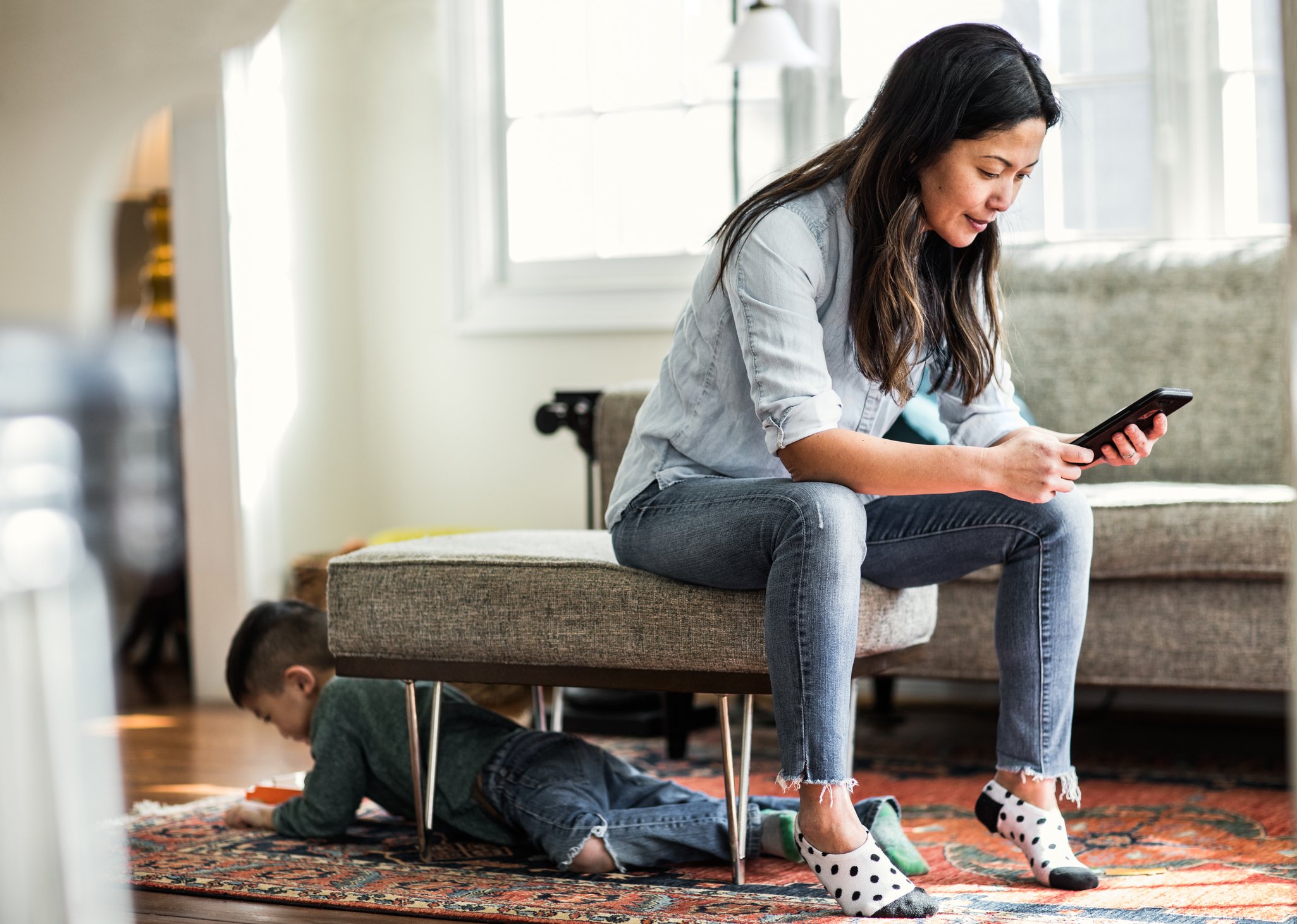 A mother scrolls on her smartphone while her child plays nearby.