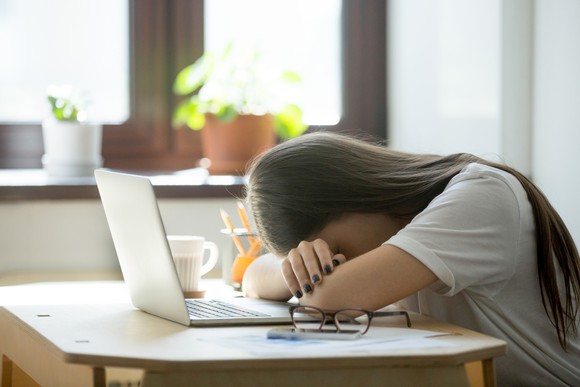 A person sitting at a table in front of a laptop computer with their head down sleeping.