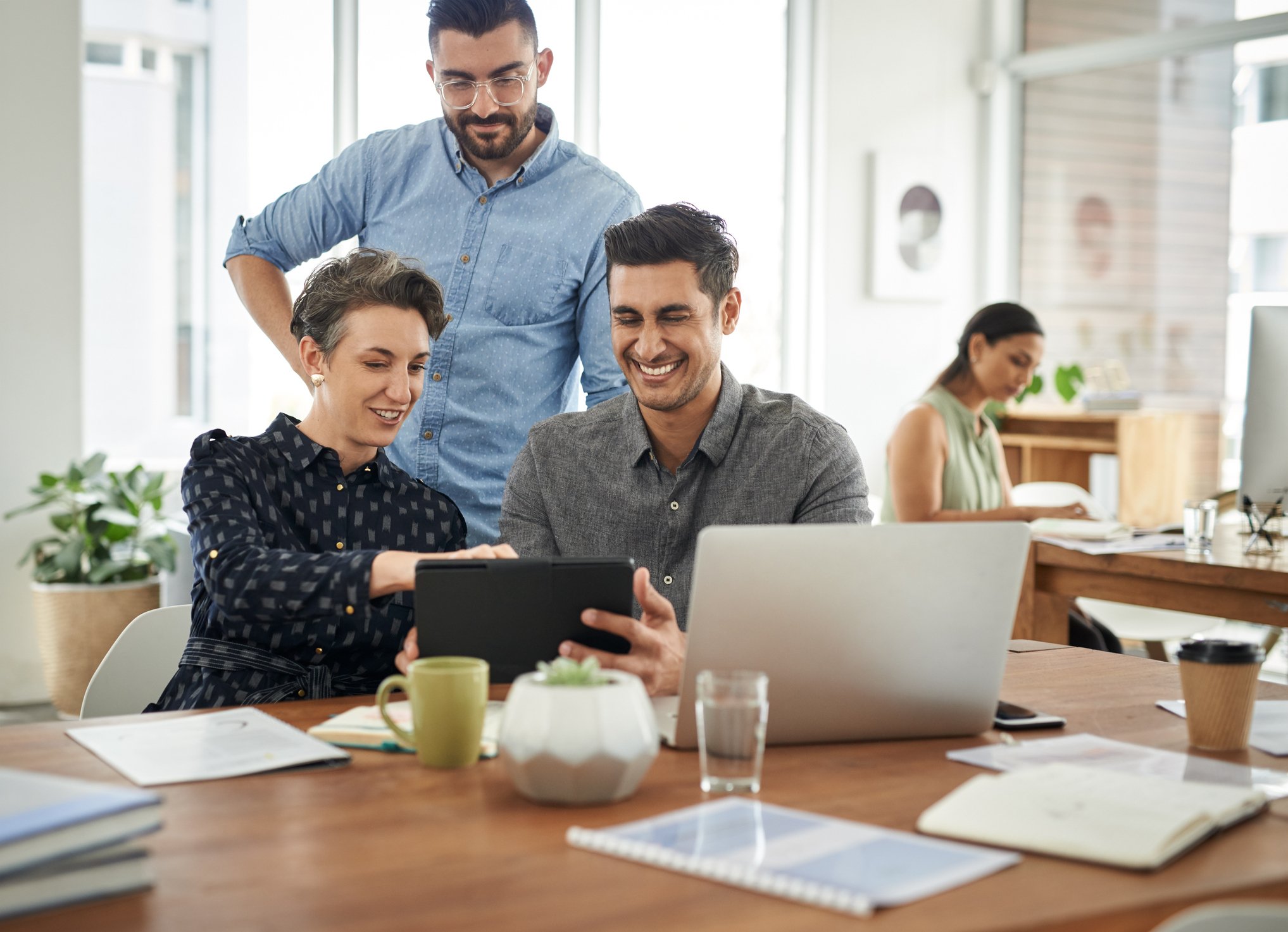 Three people smile as they look at something on a tablet in a modern office setting.