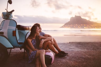 A couple at the beach leans against a scooter.