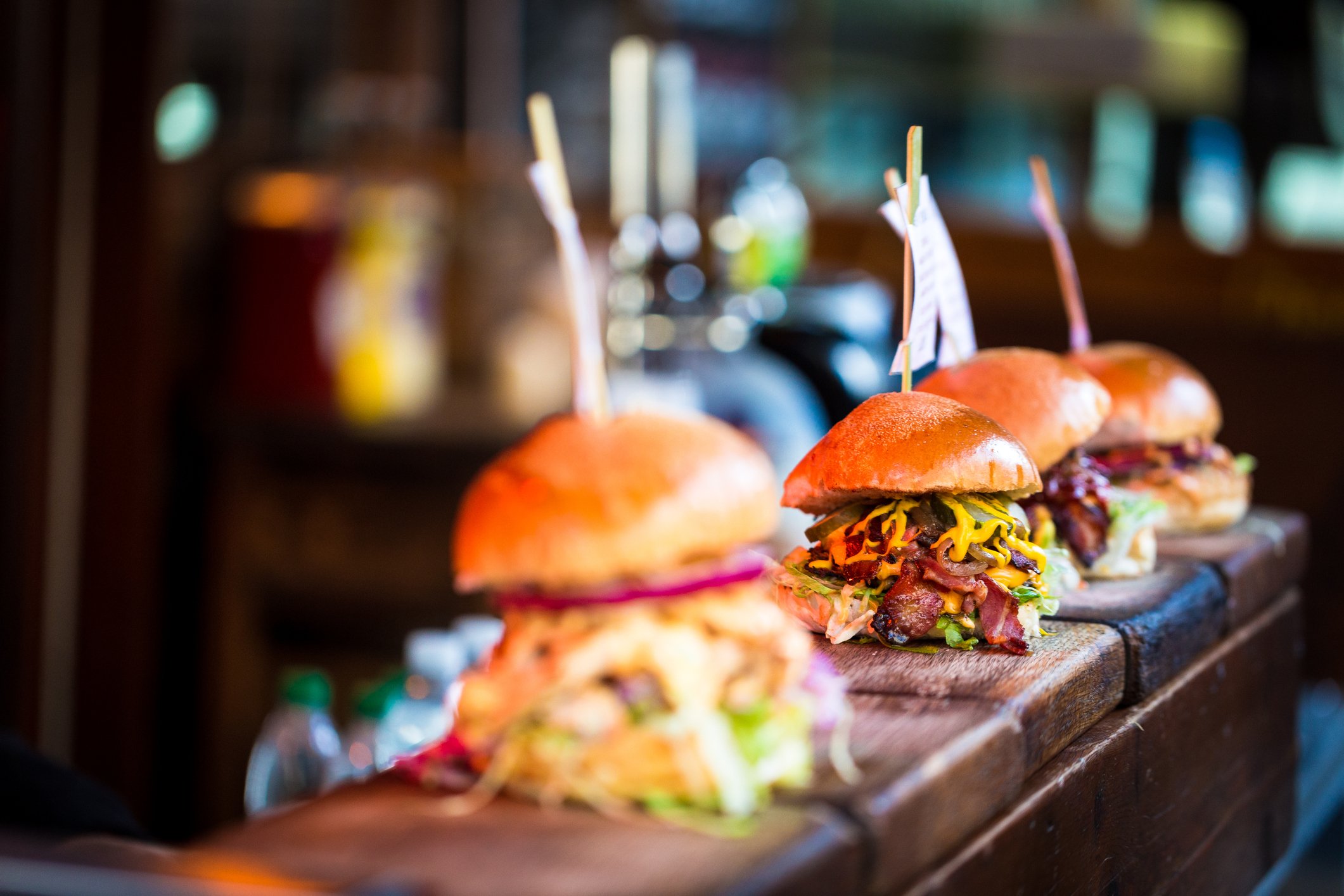 Four burgers heaped with toppings lined up on a restaurant counter. 