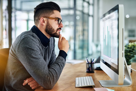 An investor strokes their chin pensively while considering a data readout displayed on a large computer screen.