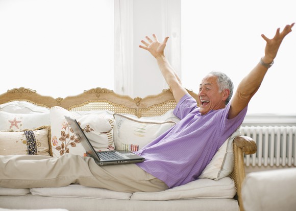 Smiling person lying on a sofa with hands in the air looking at a laptop.