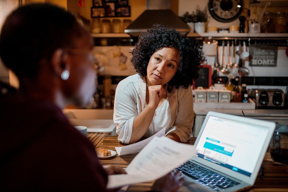 One woman at a laptop, holding papers, crunching numbers while a friend looks on, concerned. 