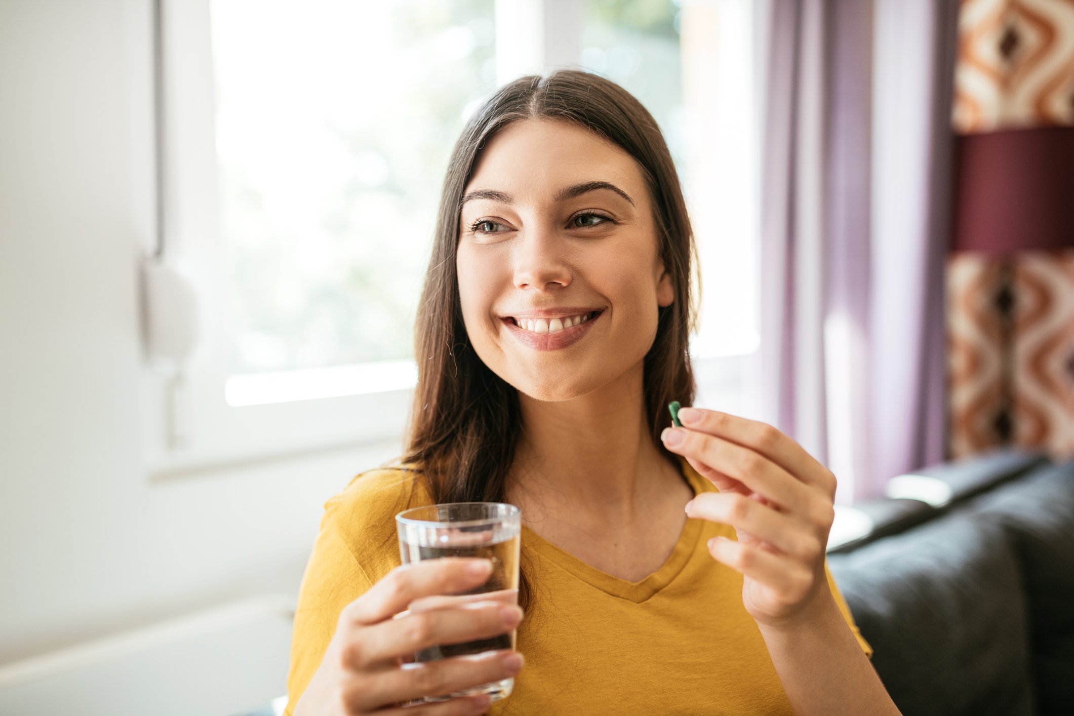 A smiling person holding a glass of water in one hand and a pill in the other.