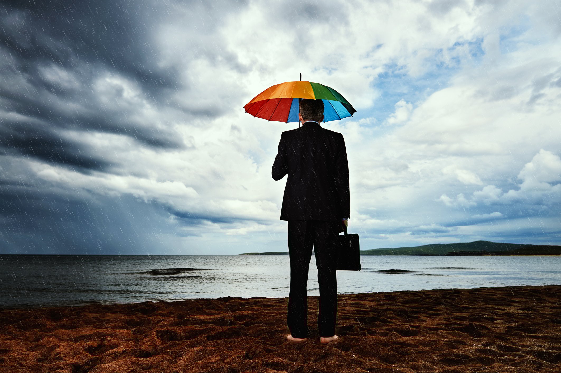 Businessperson standing in front of lake barefoot and holding umbrella as storm clouds gather in the distance. 