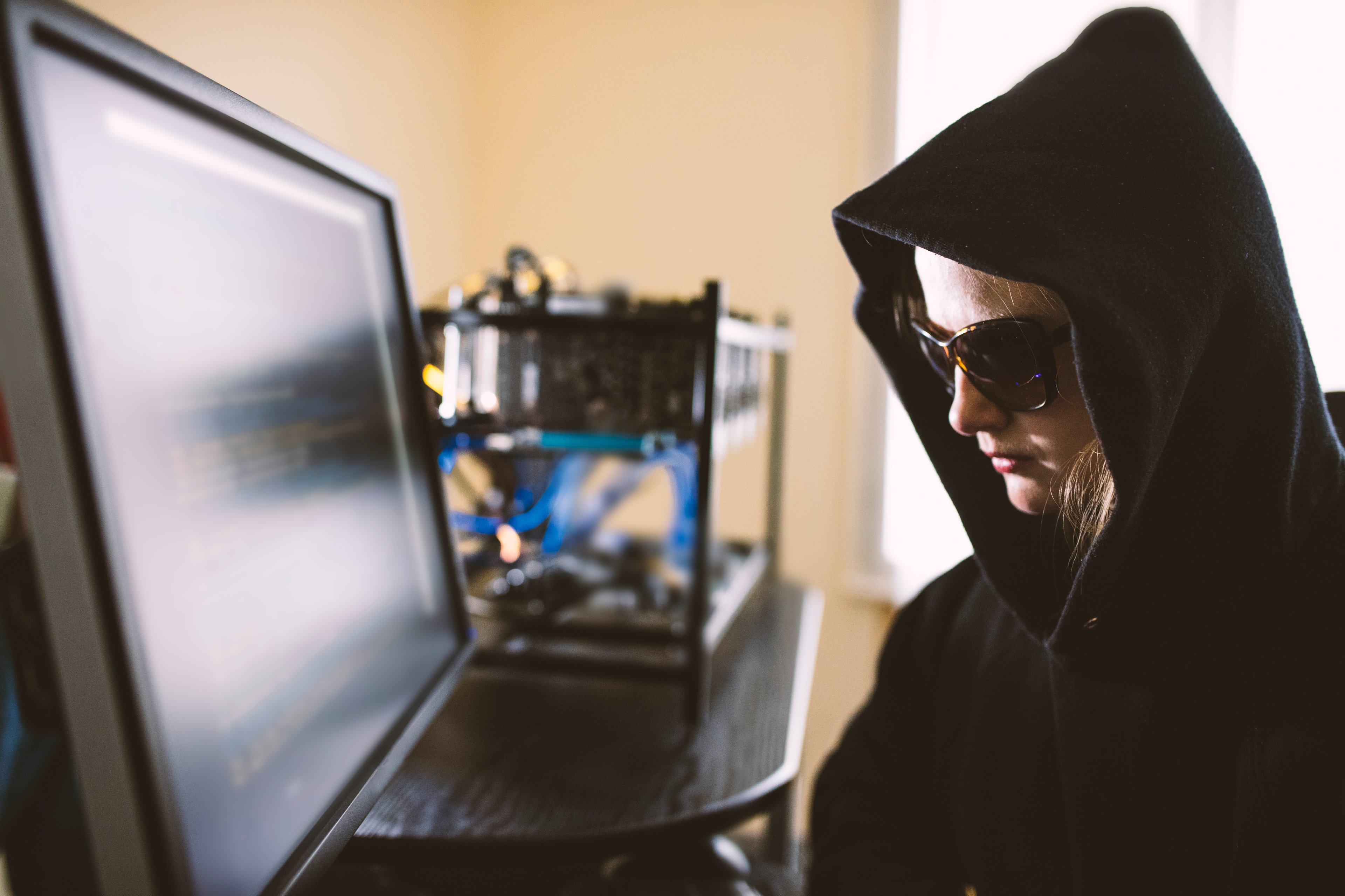 Person in black hood and sunglasses looking at a computer screen.
