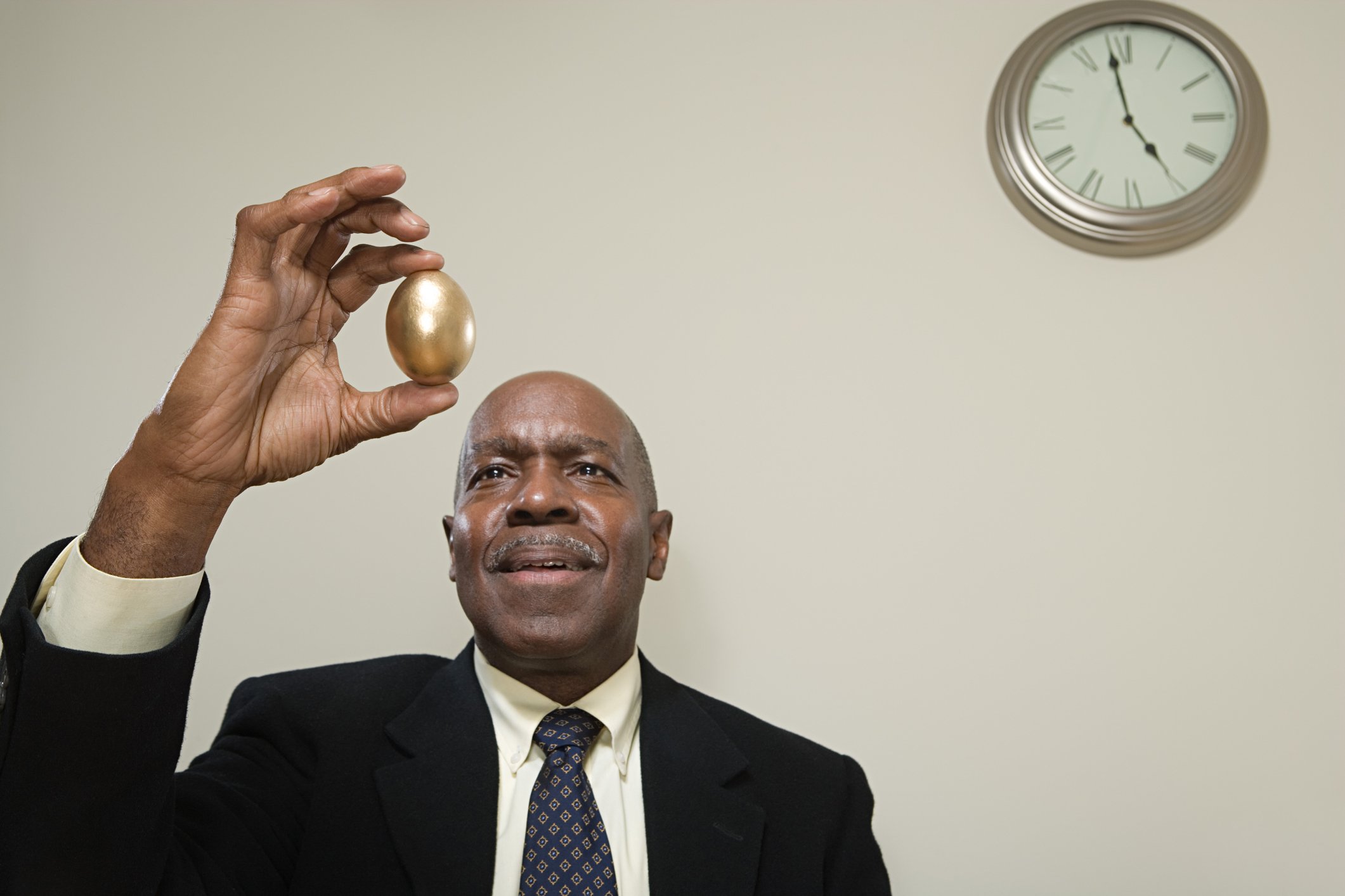 A man in a suit holding up a golden egg with a clock on the wall behind him.