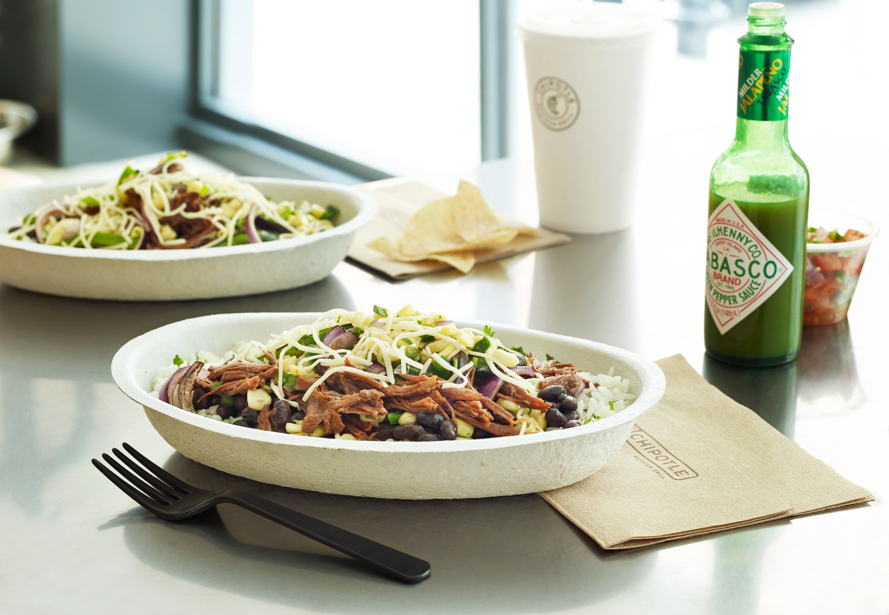 A Chipotle burrito bowl on a table next to a fork, a napkin, and a bottle of Tabasco sauce.