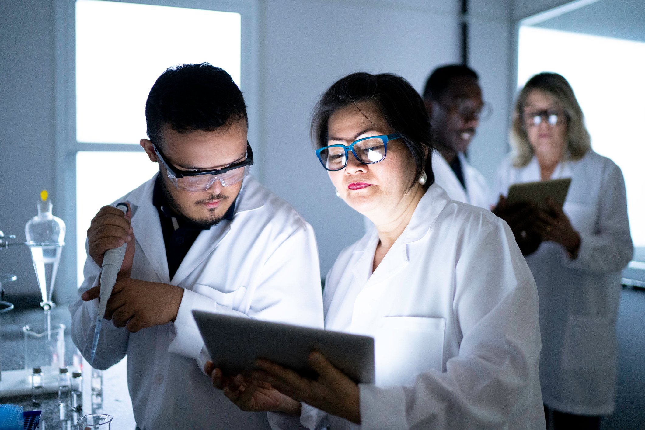 Two researchers work together in a lab, faces lit by a tablet, with others in the background.