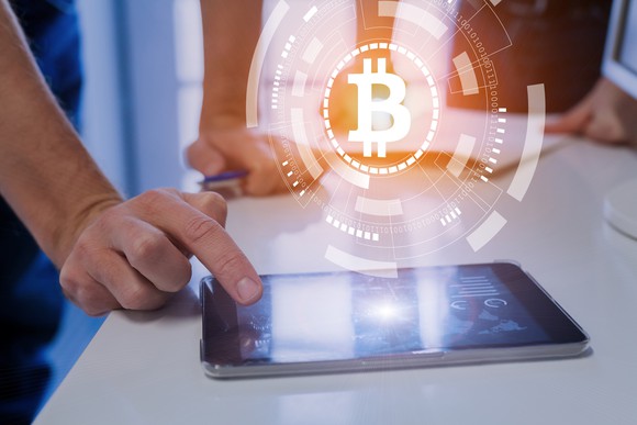 A hand touches a tablet with a bitcoin symbol superimposed over the image. 