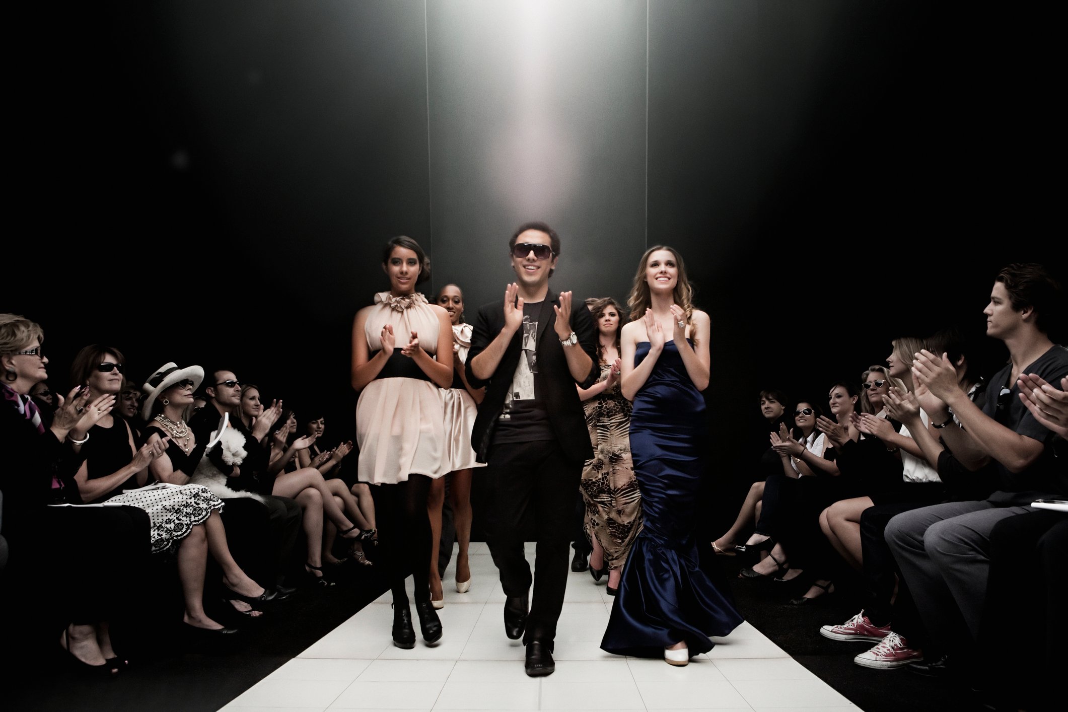 People on a fashion runway under a spotlight, with people applauding on either side.