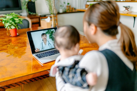 A person holding a child while looking at a laptop showing a healthcare professional.