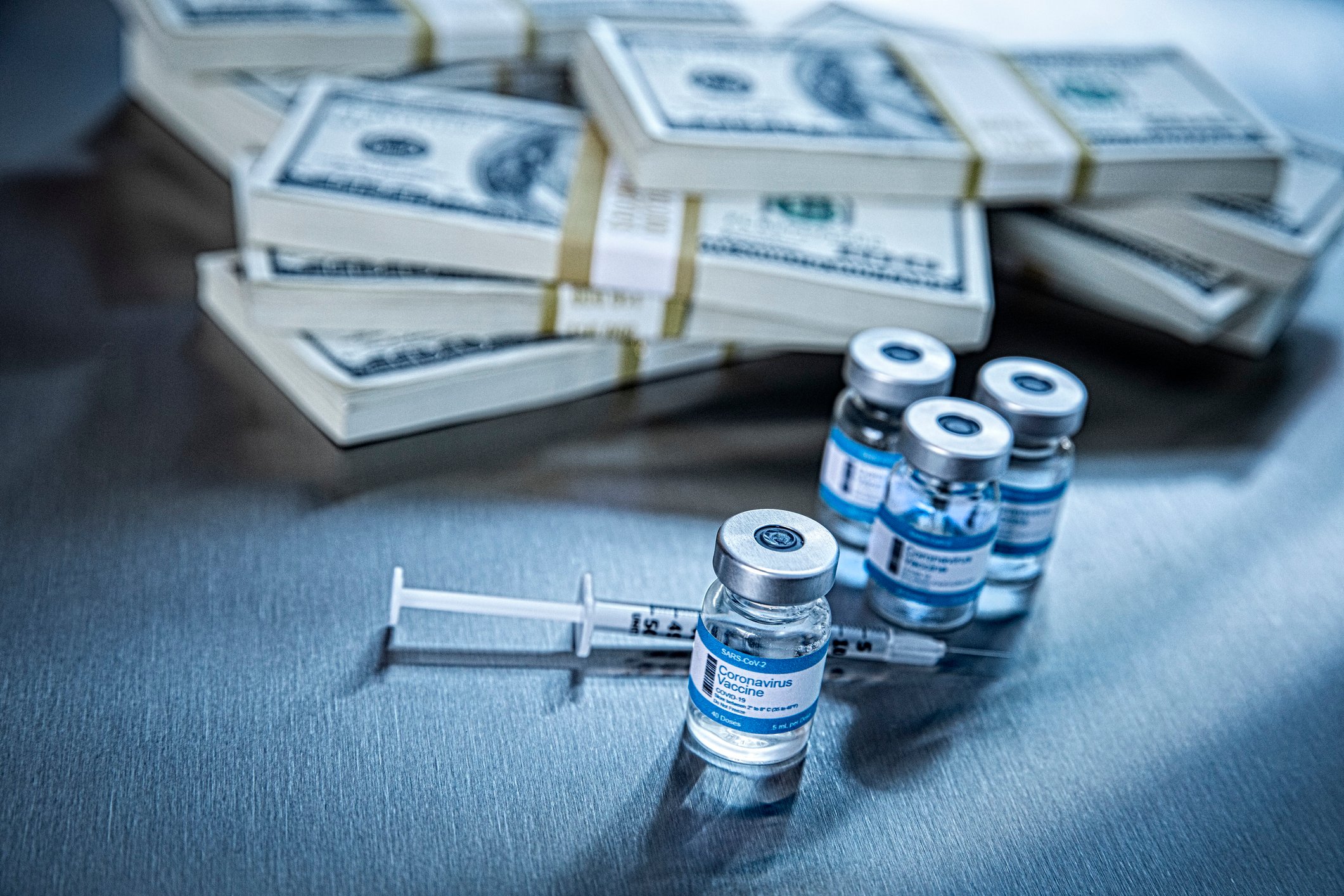 Vaccine vials with a syringe in front of stacks of $100 bills.