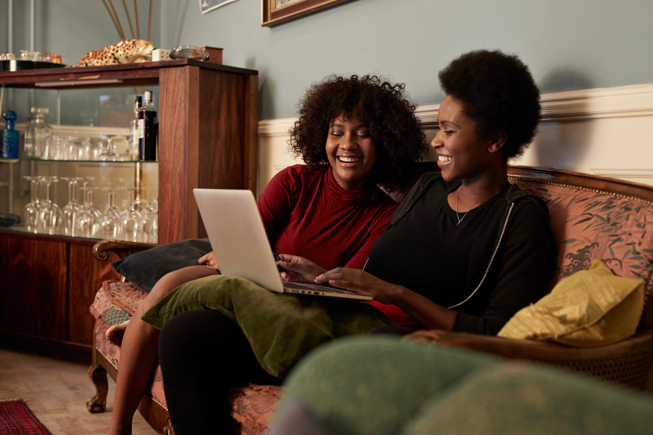 two people sit on a couch and smile while looking at a laptop.