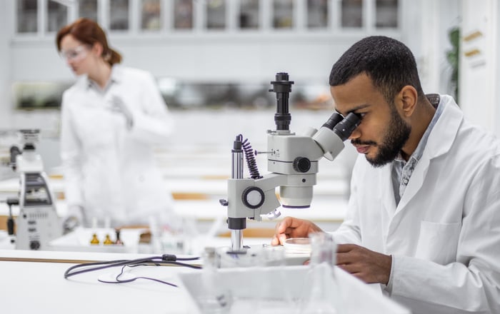 Two scientists in a lab with one looking through a microscope.